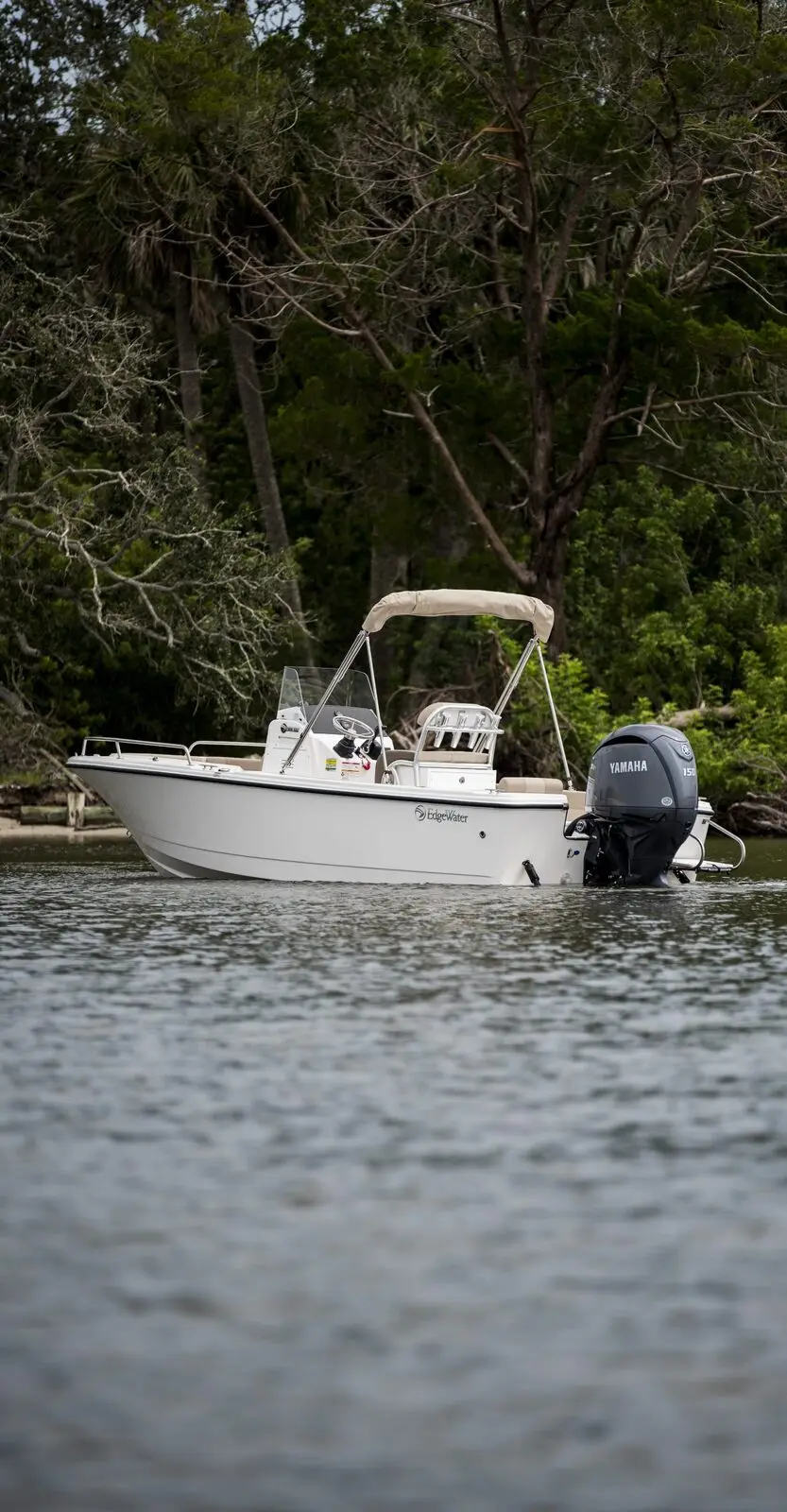 An EdgeWater center console with a tan bimini top on calm water in front of a tree line