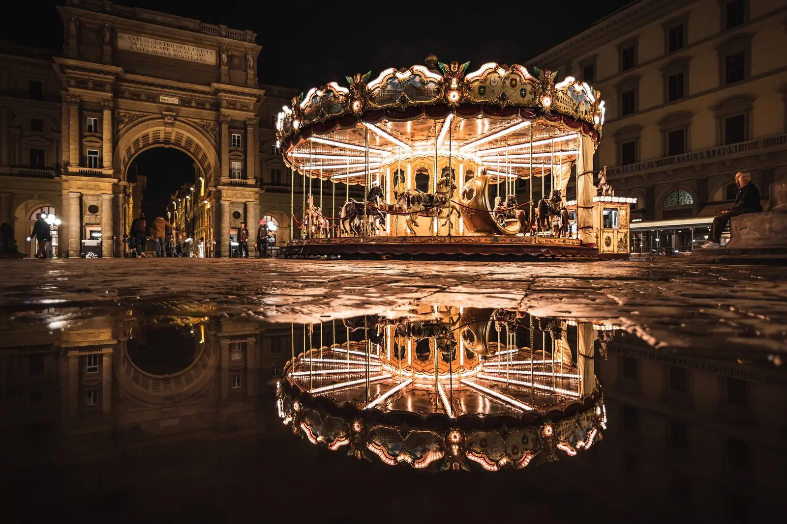 An illuminated carousel glowing at night in a city square with its reflection on wet stone, representing a small, self-contained, carefully lit world