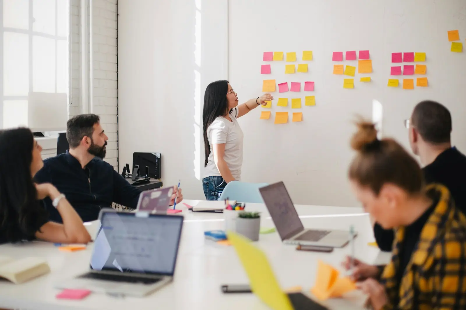 Someone arranging a wall grid of colored sticky notes while colleagues watch from a table of laptops